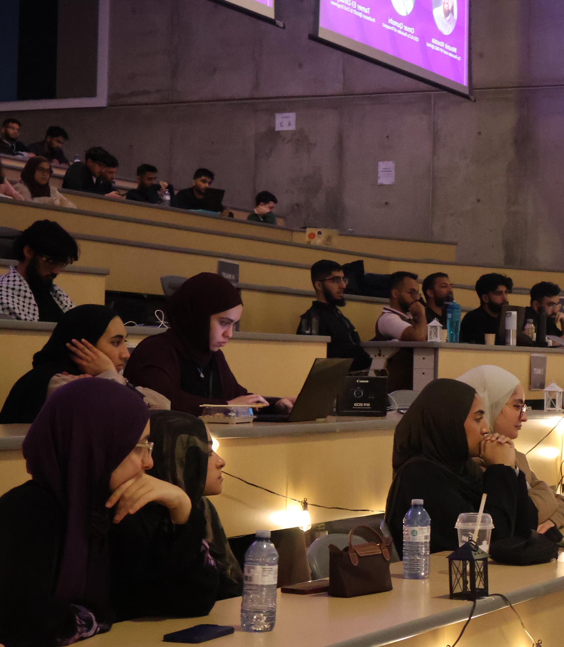 People seated in a lecture hall, with some actively taking notes on laptops and others listening. Tables are decorated with small lights and lanterns.