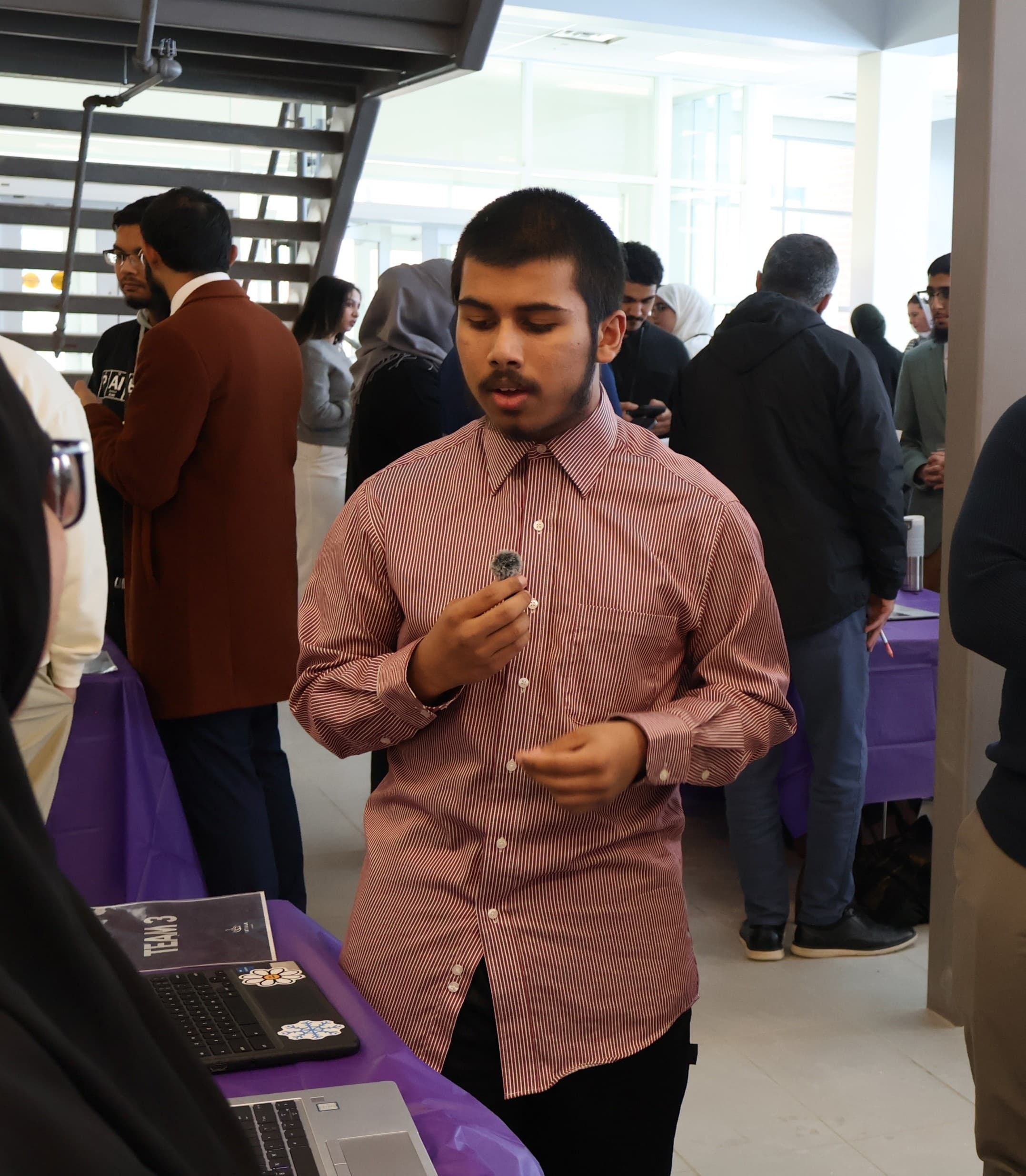 A team gives a pitch in front of two laptops at a crowded indoor event. People stand and converse in the background near a staircase.