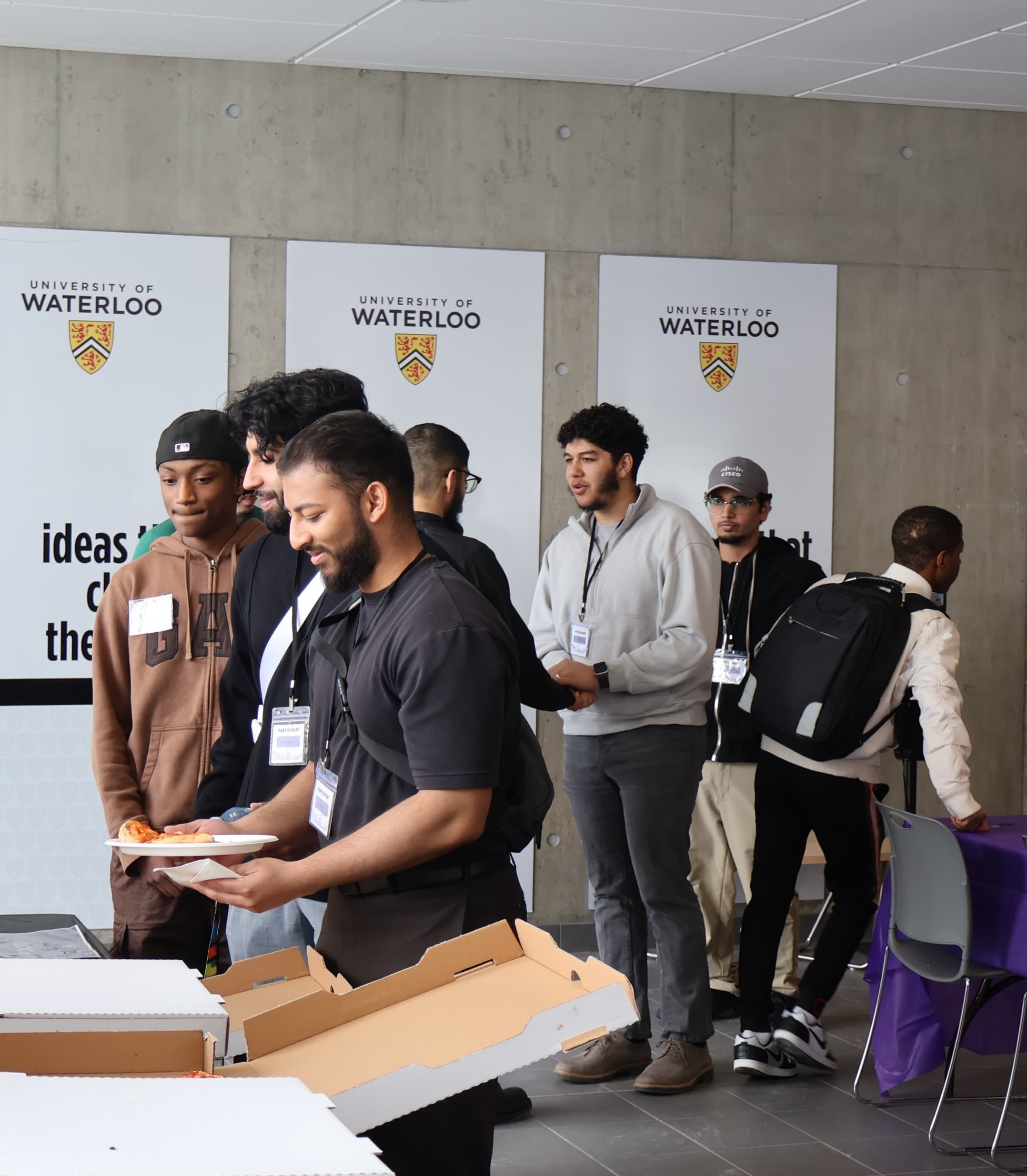 Several people of various backgrounds line up before a table to get pizza and beverages for lunch.