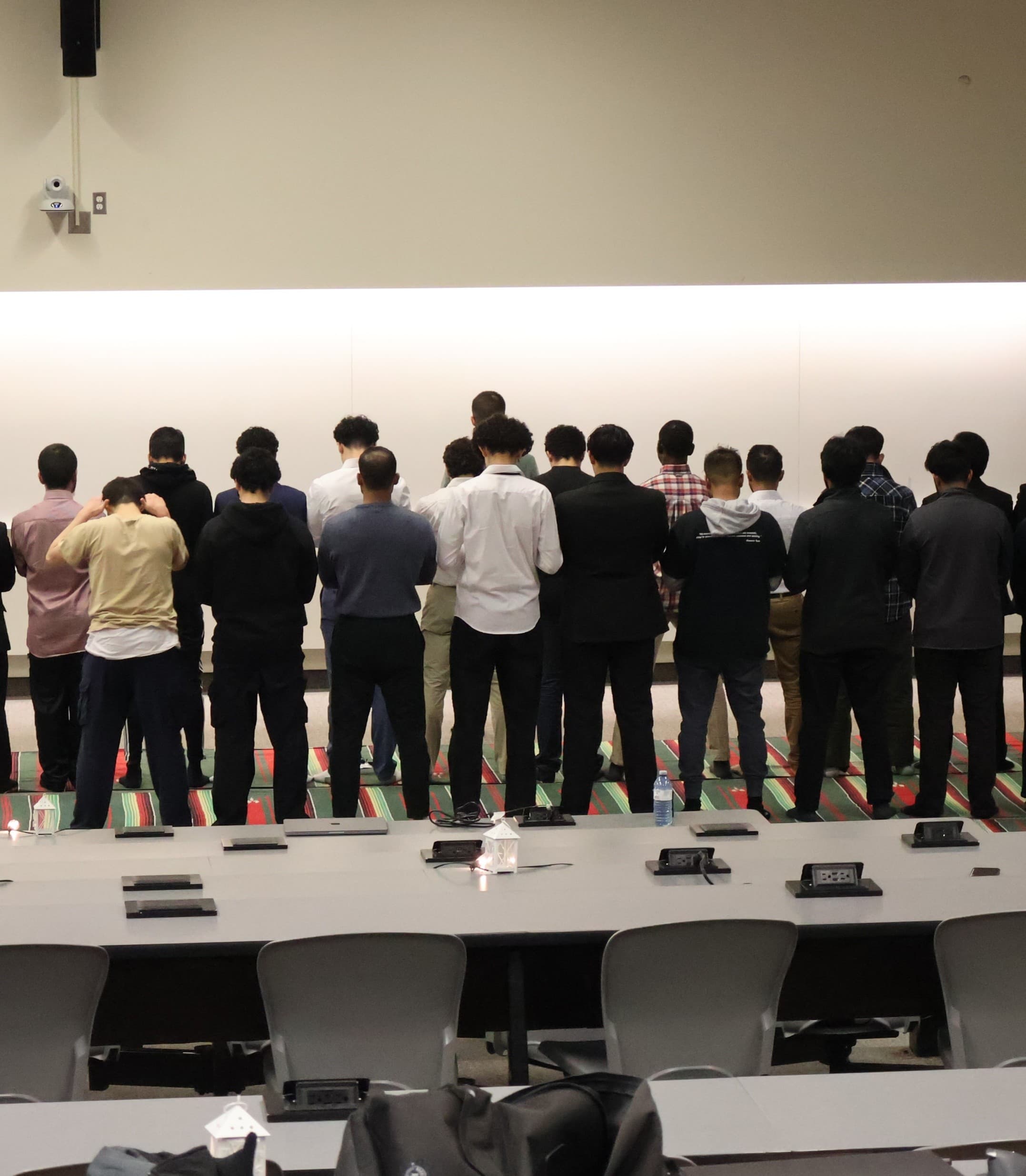 One man leads a congregational prayer, with others standing in multiple parallel rows behind him.
